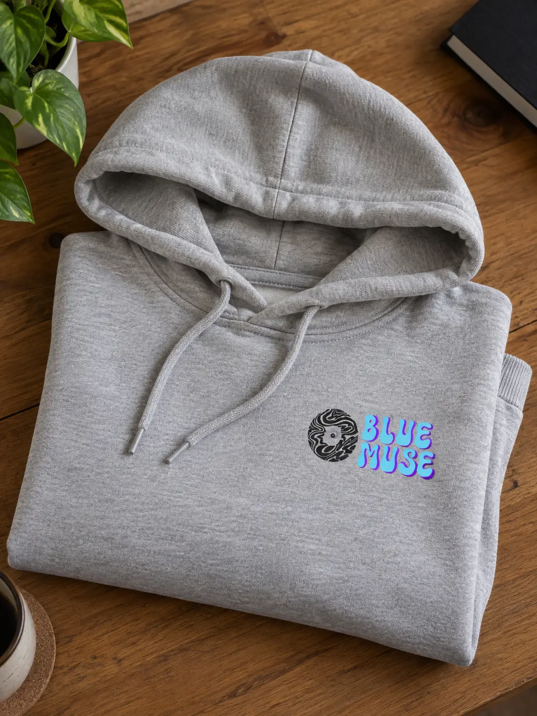 A folded heather-grey hoodie on a wooden table with the BlueMuse logo printed small on the left chest.