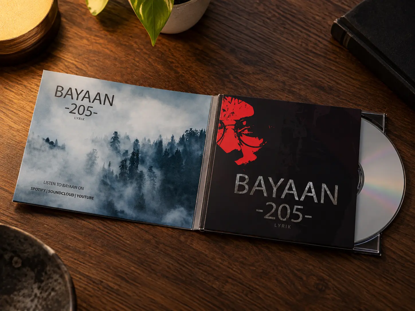 The Bayaan CD digipack open on a dark walnut table: the red-faced cover on the front, the misty forest inside, and the silver disc half out of its tray.