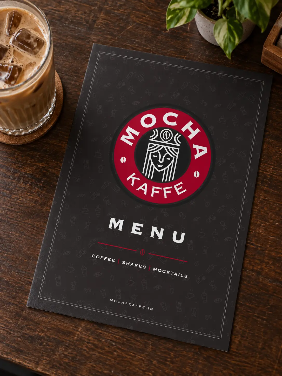 The Mocha Kaffe menu card, the crest on coffee brown, lying on a wooden table next to a glass of cold coffee.