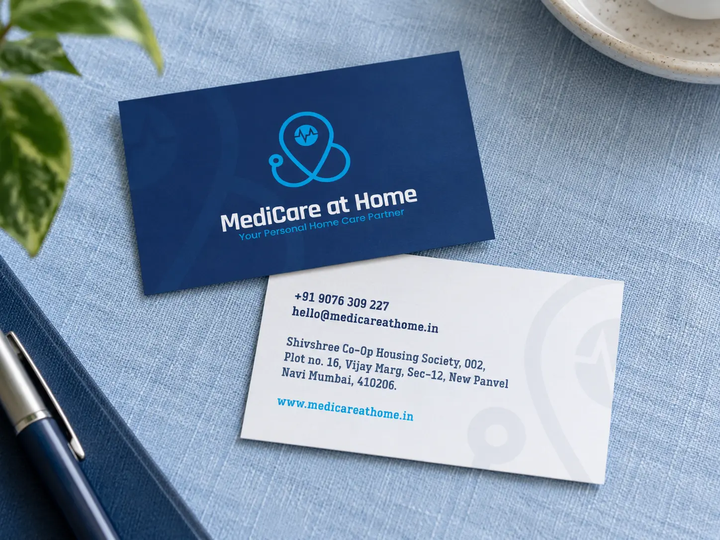 Two MediCare at Home business cards on pale blue linen: the navy front with the logo, and the white back with phone, email and address